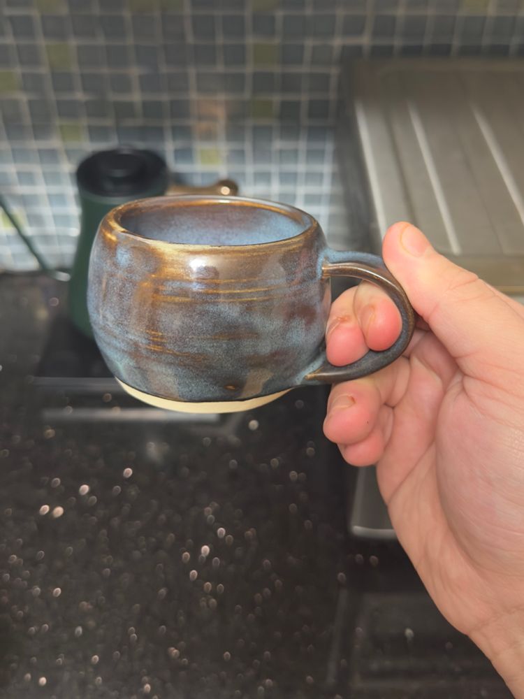 A very round handmade mug, held aloft by the handle. It’s short and stout, and glazed in a floating blue that breaks to amber over the throwing rings on the outside of the body.