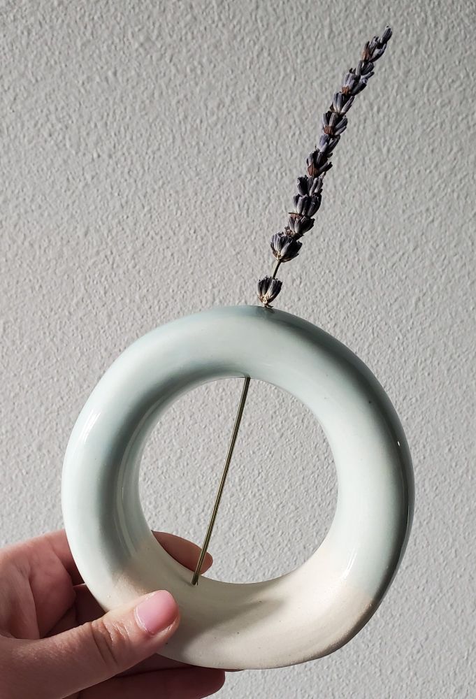 A doughnut vase with light blue glaze and a sprig of lavender. 