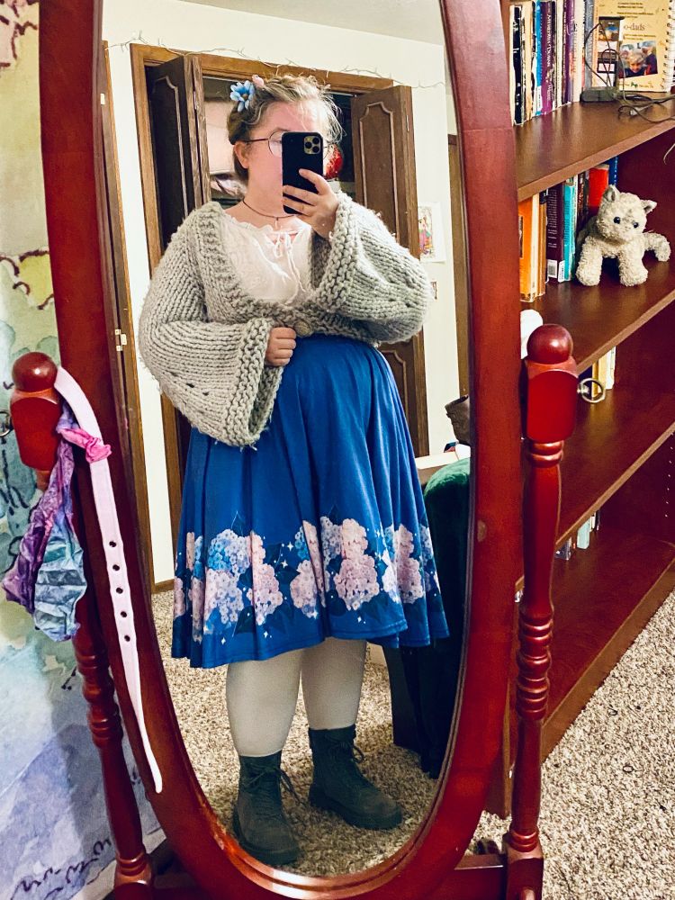 A full body mirror selfie. I am wearing a hand knit gray cropped cardigan with a wide neckline and bell sleeves. Under the cardigan is a white blouse tucked into a blue calf length circle skirt, which has pastel blue purple and pink hydrangeas around the bottom. The outfit is finished with white tights and gray boots. My dishwater blonde hair has been braided and pinned to the top of my head like a crown, a blue flower in it. My face is obscured by a cellphone and I’m wearing an iridescent crystal pendant. 