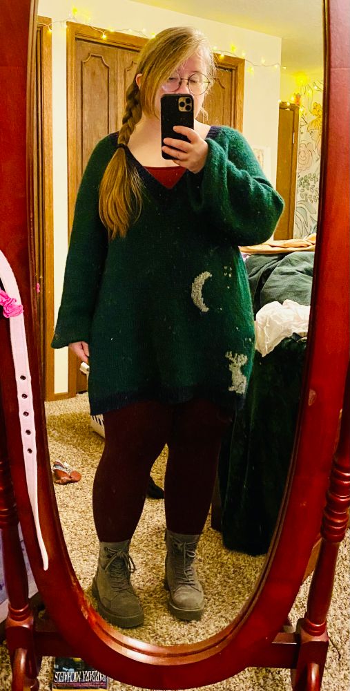Short blond woman wearing tunic length green sweater, maroon tights and gray boots. The sweater design includes a gray moon, jackalope, and stars on the bottom left. 