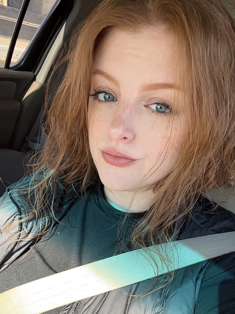 Selfie of ems in a car with tan interior . They are wearing their hair down , a light face of makeup , and a black outfit . 