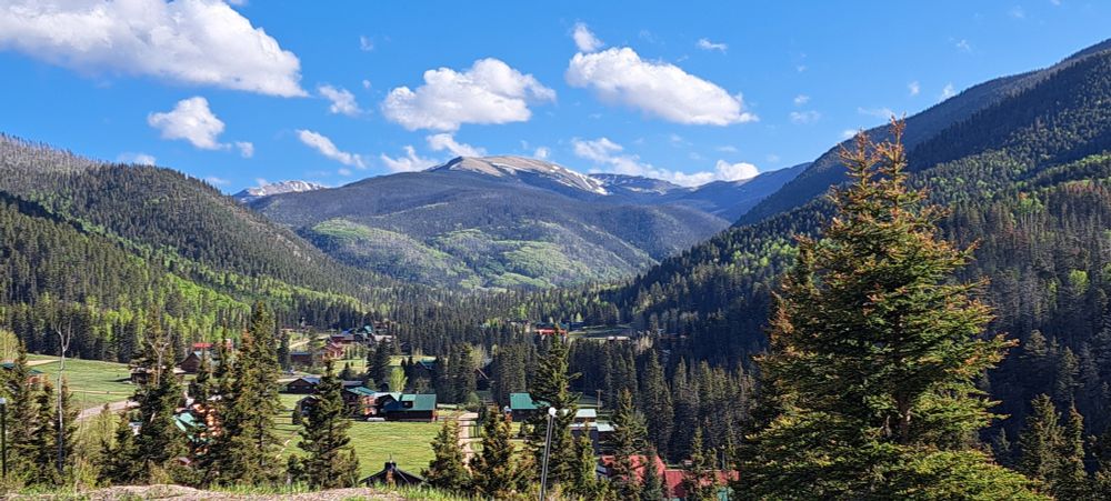 Northern New Mexico, Upper Red River Valley, early June. Frazier Ridge with Wheeler Peak behind. Conifers, aspens and grass provide shades of green. There is snow on the highest elevations. The sky is blue with some puffy white clouds.