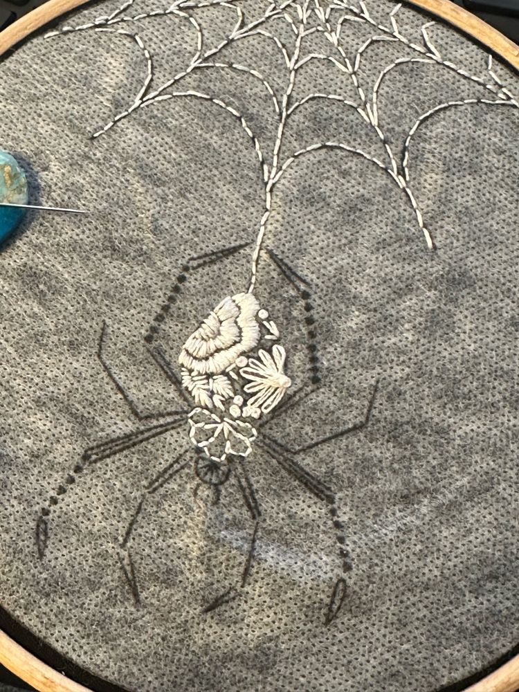 Black cloth with soluble gray stabilizer in an embroidery hoop. On the cloth is incomplete embroidery in white thread depicting an Orb Weaver spider descending from a bit of web.