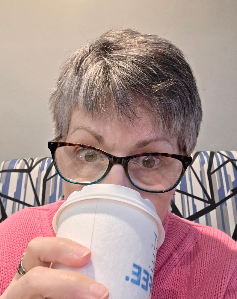 A gray haired white woman slugging down hotel coffee and contemplating never traveling by car again when it's below freezing outside. 