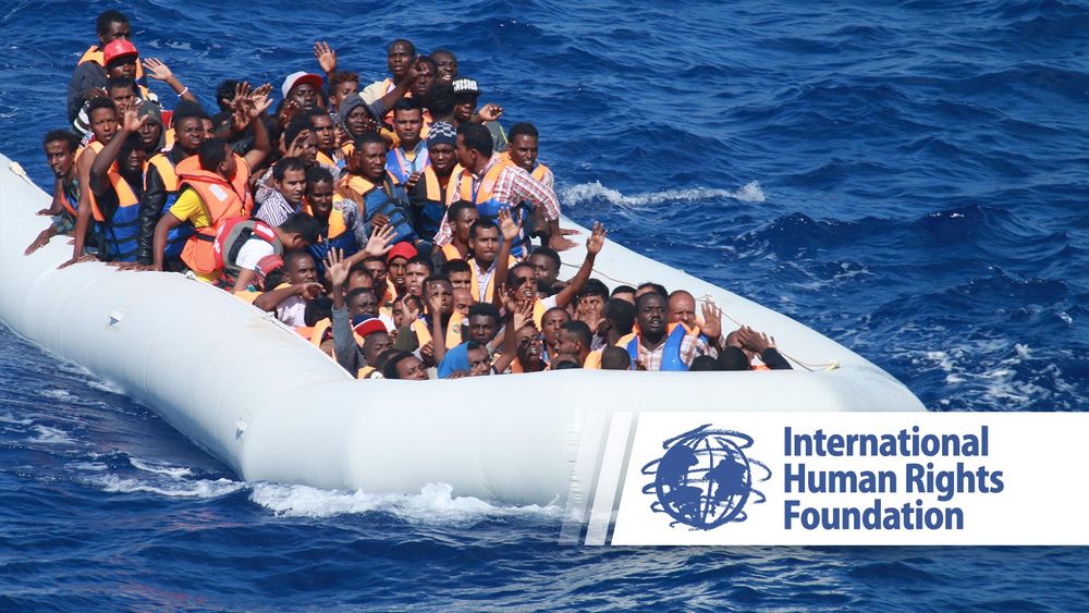 The image shows an inflatable boat overloaded with a large number of people, many of them wearing life jackets, sailing on the open sea. The people on board appear to be migrants or refugees, and several of them are raising their hands, possibly asking for help or waving. The sea is a deep blue colour and there are no other boats in the vicinity. In the lower right part of the image, there is a logo and the name of the organisation ‘International Human Rights Foundation’, which suggests that the image is related to humanitarian activities or raising awareness about the migration crisis.