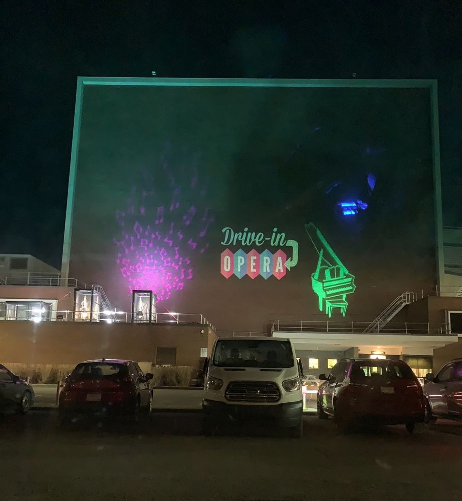 this was during the pandemic lockdown. a photo taken from sitting in my car, in front of a building at night. there are projections on the building of music notes and a piano and the words “drive-in opera”.