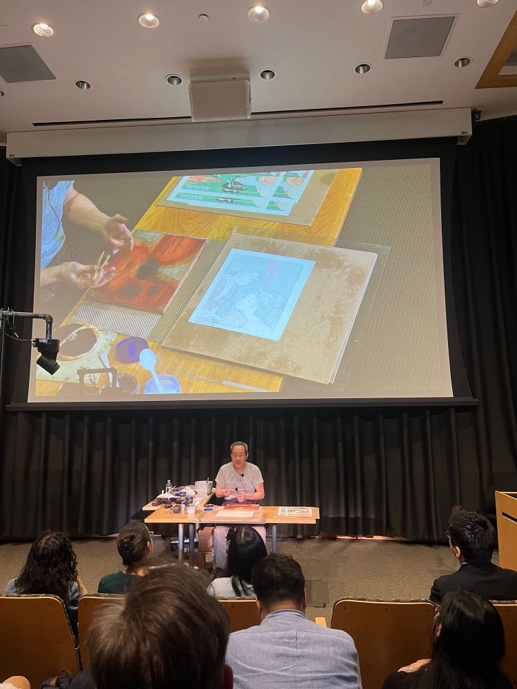 Man at a table with tools, paper, inks. Behind him is a screen showing a close up of the items on the table, including a woodblock with a deep orange-red ink