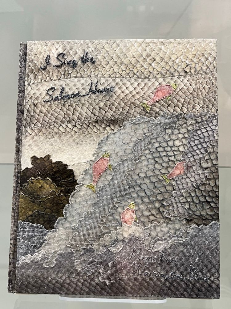 Book cover with scales and dyed sections creating an image of blueish water, jumping pink salmon, and grey sky