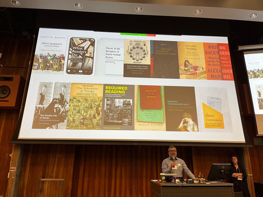 Screen of 13 book covers with presenter with mic speaking below the screen