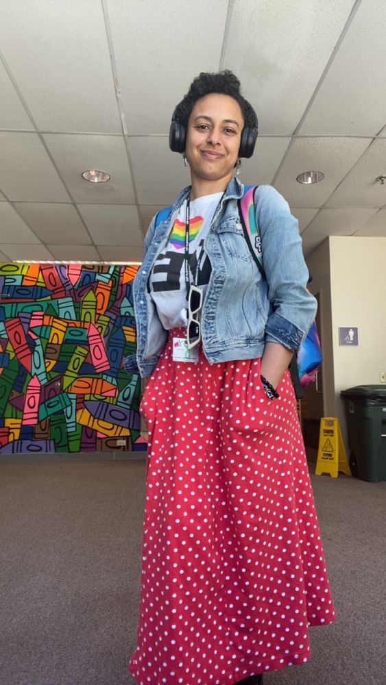 Taylor wearing black stereo headphones standing in front of a mural of crayons and public bathrooms. They are wearing a jean jacket, white shirt with the word “LOVE” on it with a rainbow heart, and a long red and white polka dot skirt with pockets! They have a multicolored backpack on.
