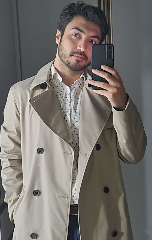 Pessoa tirando uma selfie no espelho, vestindo um trench coat bege sobre uma camisa branca com pequenos padrões escuros. A pessoa está segurando um smartphone, cobrindo o rosto com ele para tirar a foto.