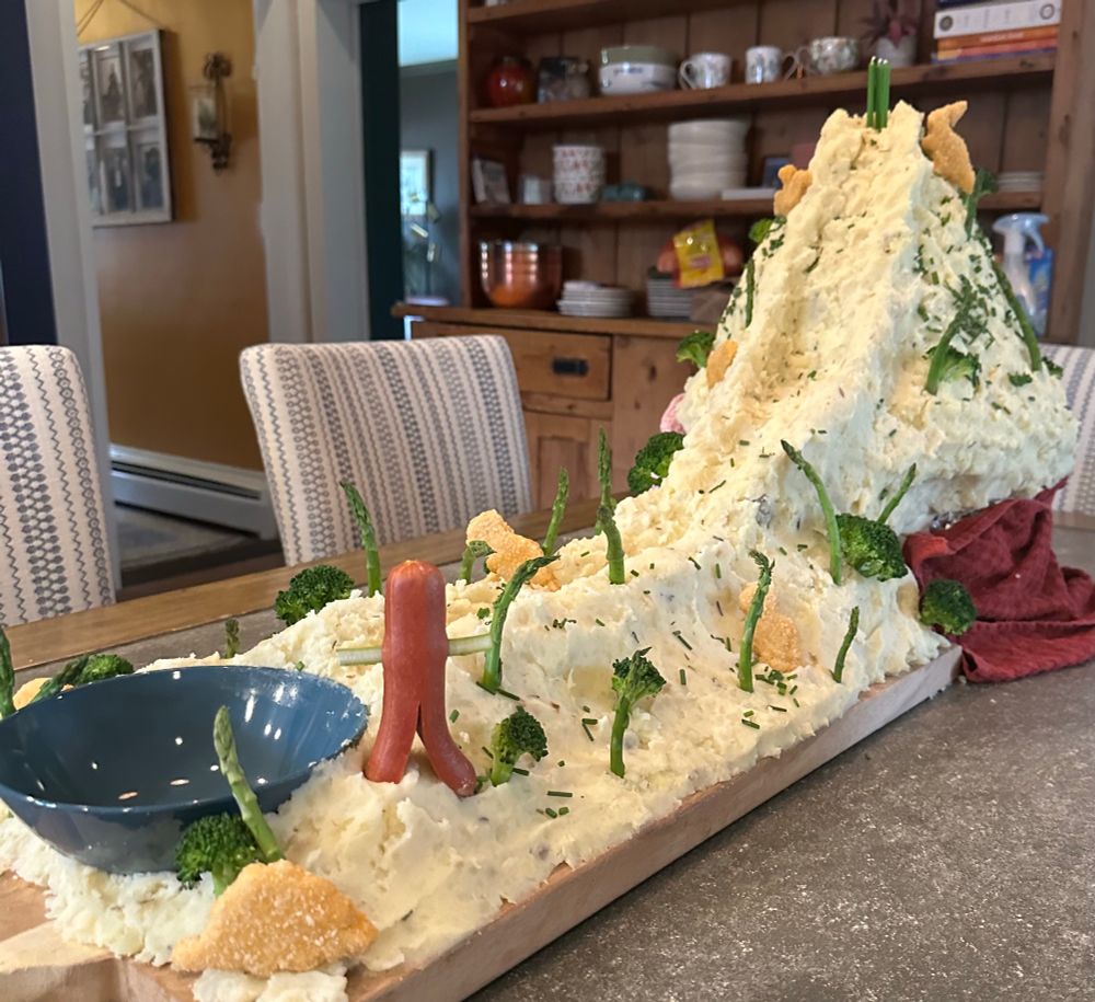 A volcano made out of mashed potatoes with asparagus and broccoli trees mingled around Dino nuggets and a hot dog shaped like a person. 