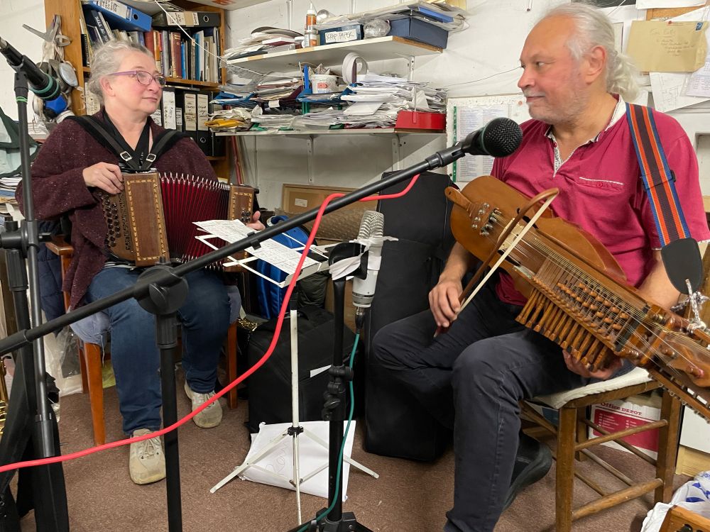 Heather playing melodeon + Ravi playing nykelharpa sit in a small office space. There are a few microphones and stands dotted around. 