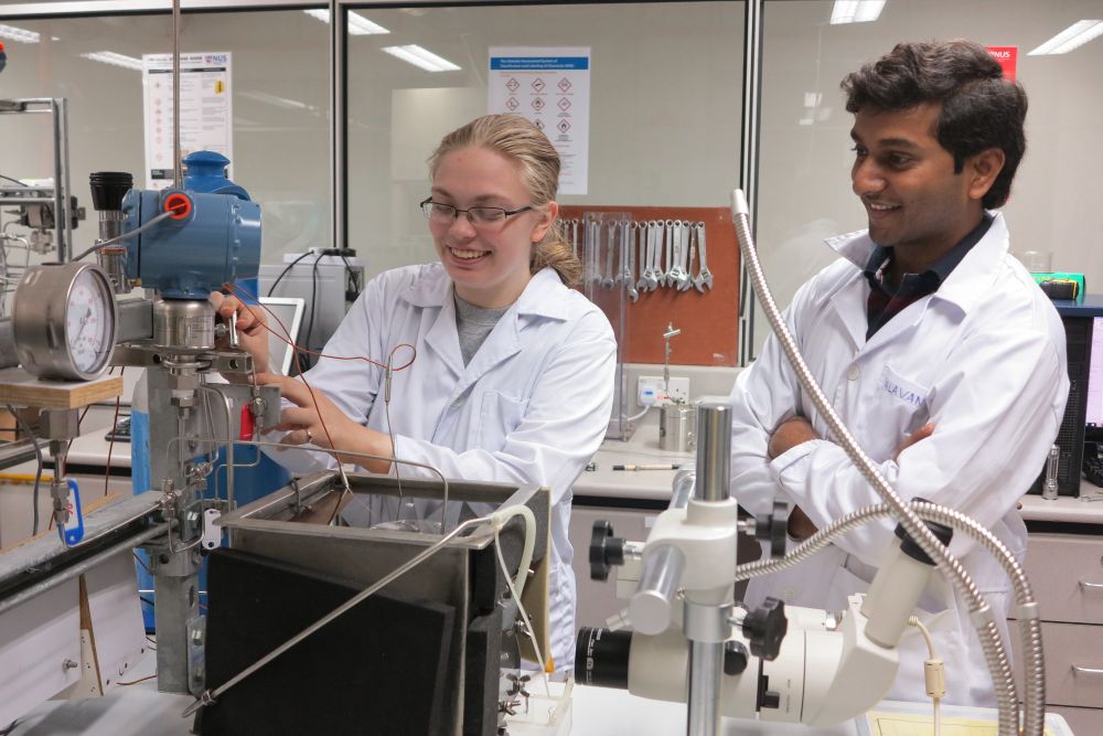 Jessie, an undergrad, stands next to Valavan, a grad student, in a chemical engineering lab. They both wear lab coats. Jessie is changing settings on a lab setup for forming gas hydrates while Valavan supervises.