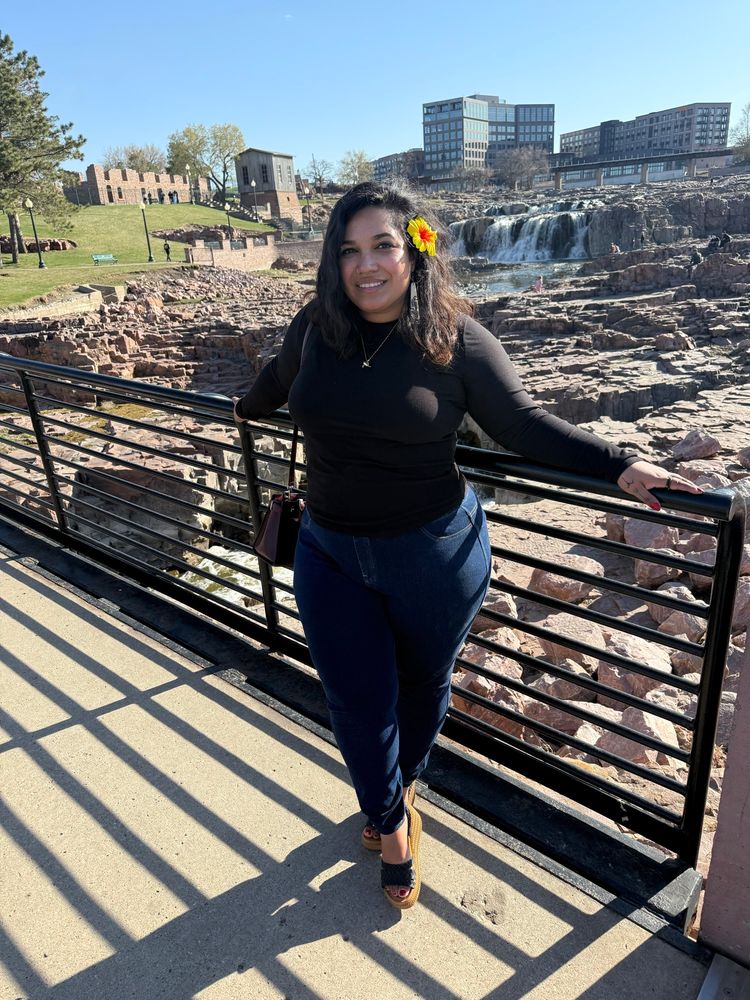 Picture of me in a black long sleeve shirt, dark blue skinny jeans, black and brown wedges, with a maroon Kate spade purse. Standing on the bridge at the falls in SD. 