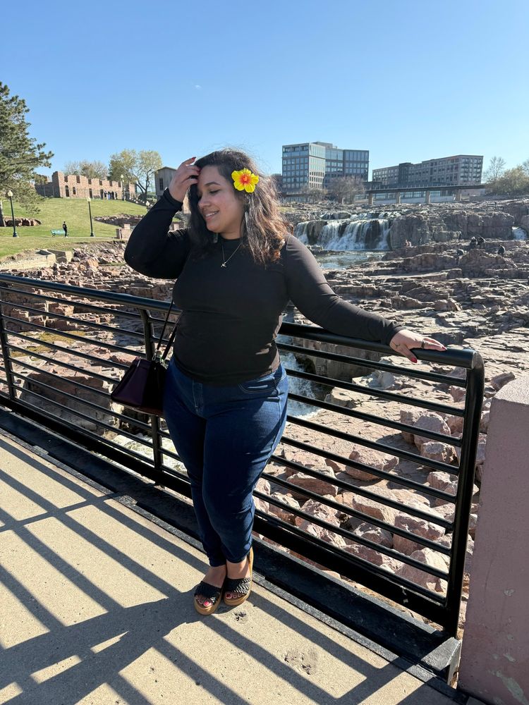 Picture of me in a black long sleeve shirt, dark blue skinny jeans, black and brown wedges, with a maroon Kate spade purse. Standing on the bridge at the falls in SD. 
