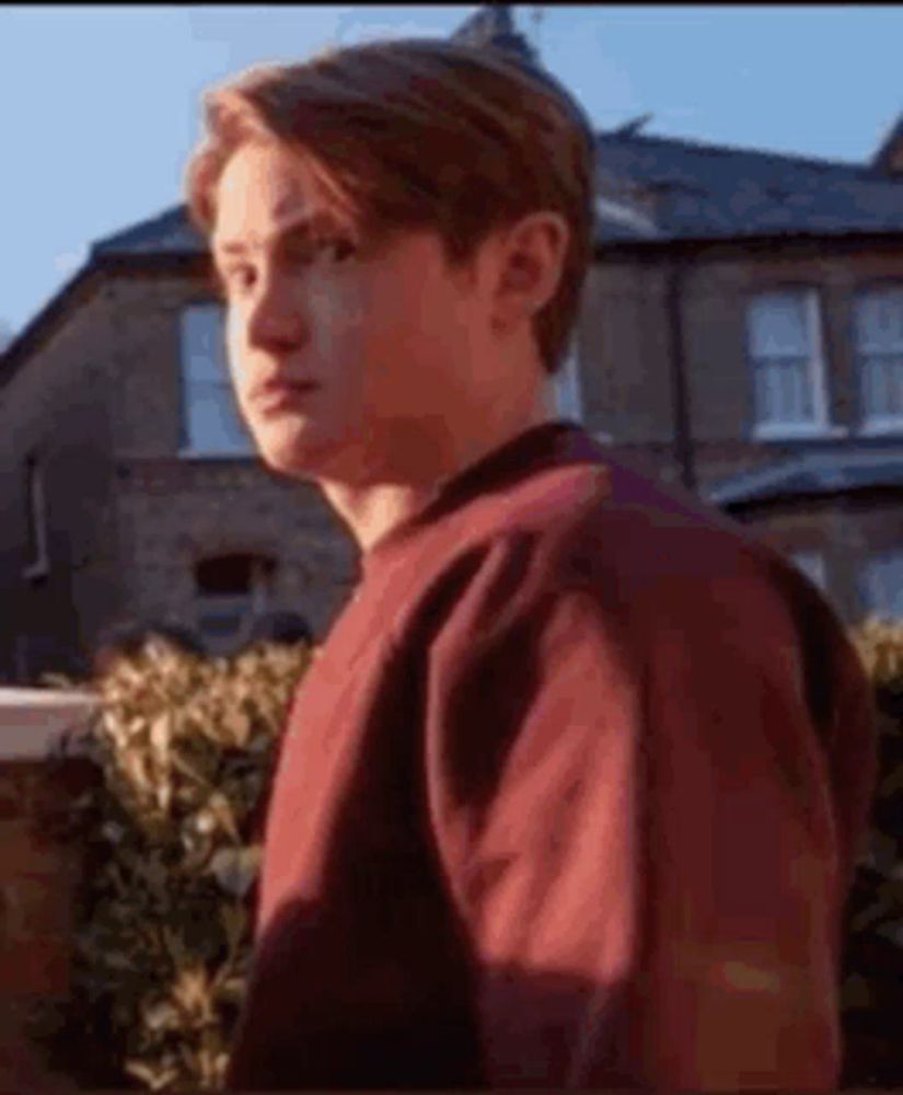 a young man in a red sweater is standing in front of a house and looking at the camera .