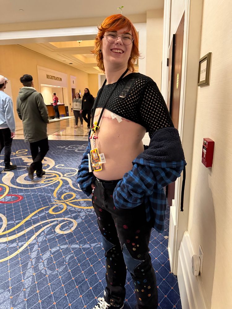 A photo of me with a goofy smile. I’m standing in the gaylord. My outfit is a skimpy knit top, my blue plaid jacket and my skinny jeans that I have embroidered swirls all over. I am also wearing converse shoes.  