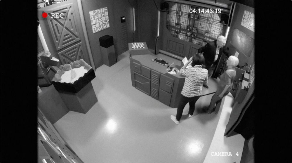 A scene from the Doctor Who Escape Room bonus feature showing members of the Doctor Who cast in an escape room styled like a 70s or 80s BBC sci fi space station 
