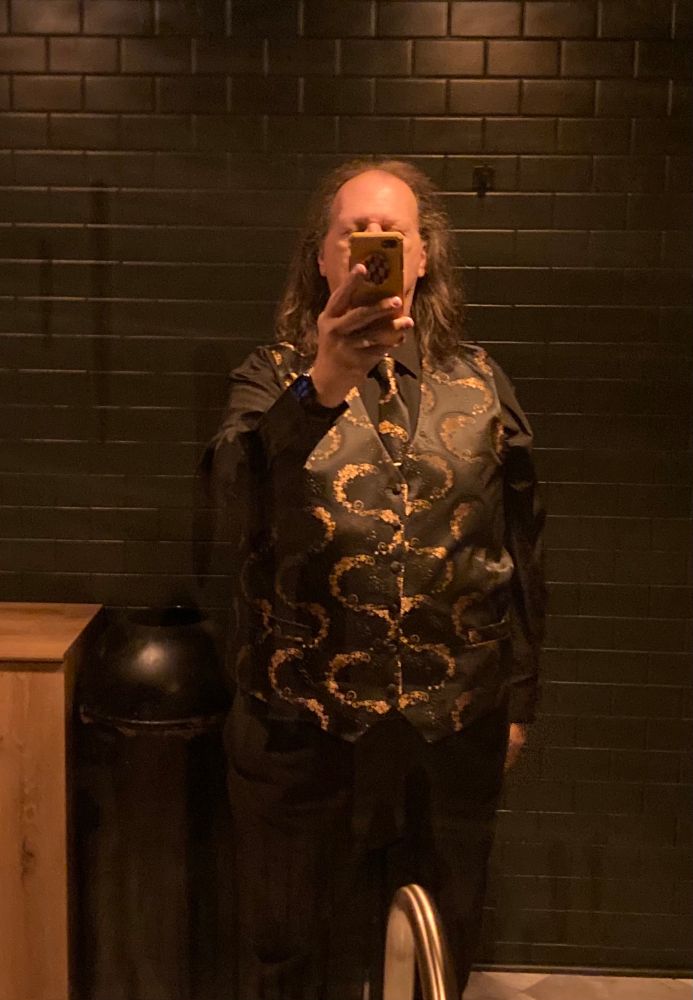 A mirror image of a man in a black shirt and black/gold vest.