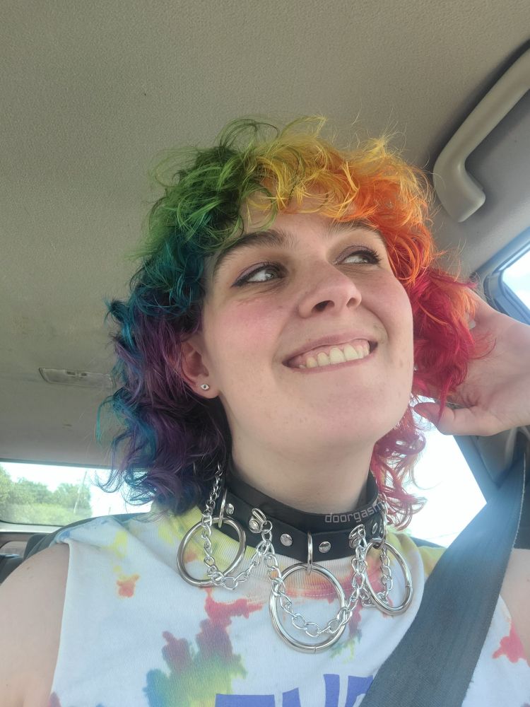 Door in their car after their hair appointment. Their hair is now a very vibrant rainbow. They have on a black choker with chains and large o-rings, and a tank top that reads "they them theirs"
