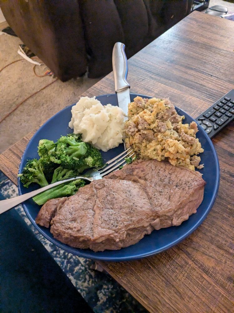 A blue dinner plate sitting on a coffee table. On the plate, from the top going clockwise, is a large scoop of sausage stuffing, a large steak, some broccoli, and a moderate scoop of mashed potatoes.