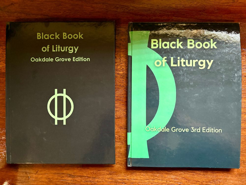 A first edition copy of Oakdale Grove's "Black Book of Liturgy" next to a third edition copy. The first edition bears a small plain Druid Sigil centered on the front cover. The Sigil and the text are lime green on a black background. The Third Edition shows half the sigil very large centered on the book's spine so that the full sigil is seen by others while the book is held open by the reader.