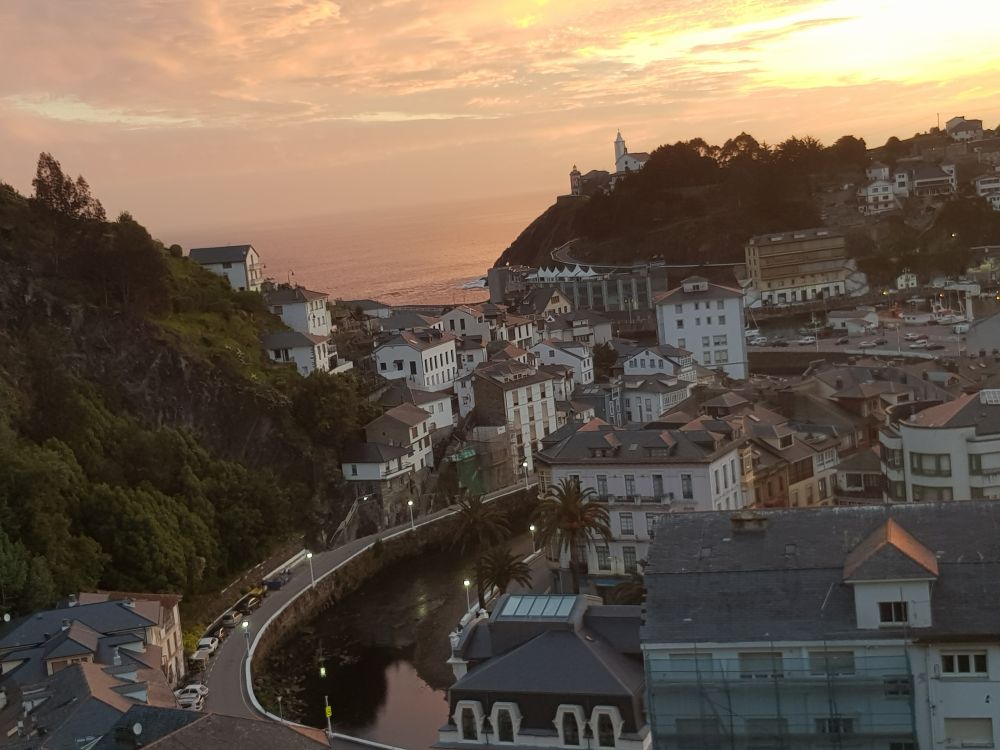 Bild från en höjd mot den lilla staden Luarca med dess huvudsakligen vita små hus. En flod kommer in från höger på bilden och böjer av uppåt, tillbaka, åt höger innan den rinner ut i havet på bildens översida. En liten kyrka på en höjd till höger vid havet 