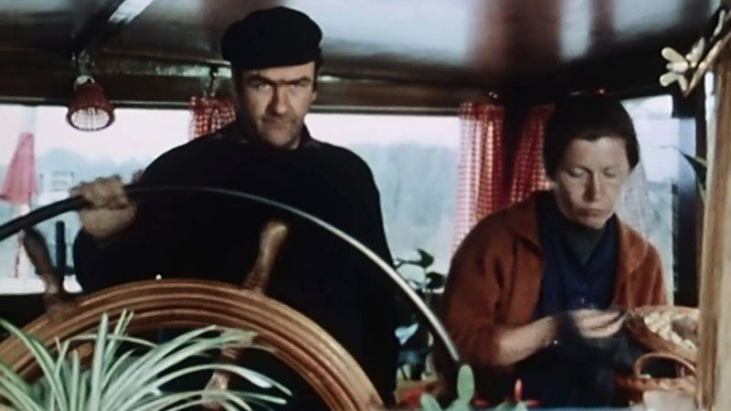 Un marinier à la barre de sa péniche avec sa femme qui cuisine à côté.