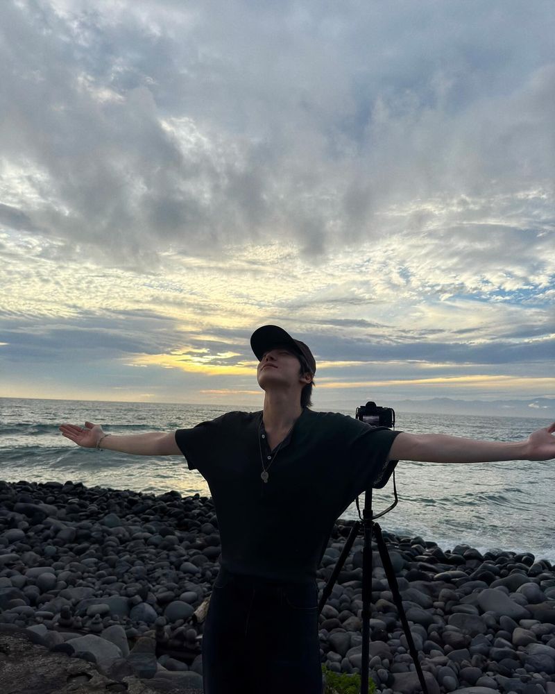 Yunho wearing a dark three button t-shirt, standing on a rocky beach in a T pose with the ocean and the cloudy sunset behind him. His DSLR is on a tripod behind him. It's like you took the photo (but it was actually Mingi). 
