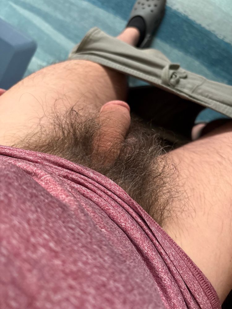 POV looking down at a man with his pants down around his ankles and a long, natural bush is showing over a large, flaccid penis