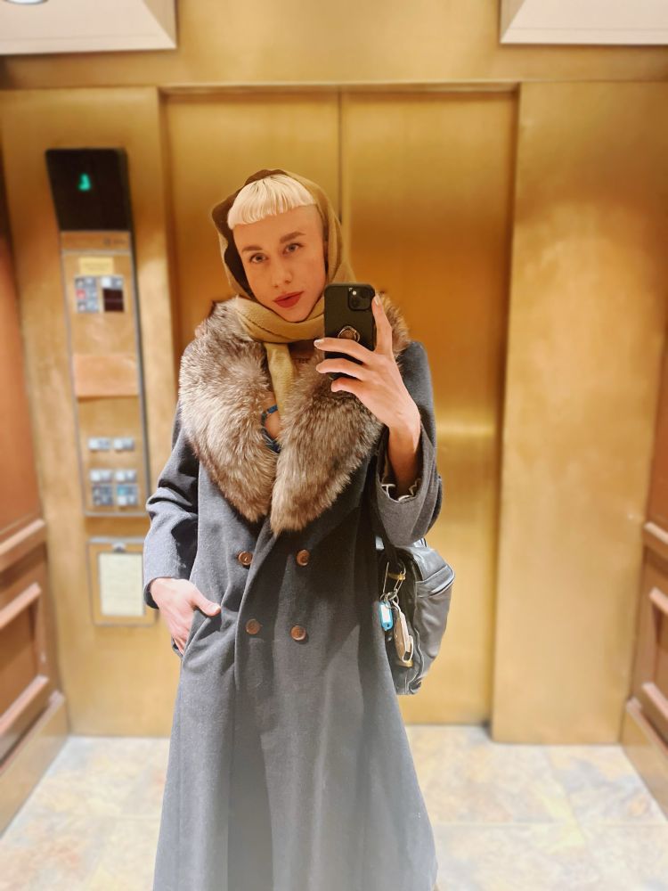 Wearing a grey wool coat with fur trim & my hand is tucked into my pocket. I am gazing into a mirrored wall, my blonde hair bright under warm light. Golden elevator doors close behind me.