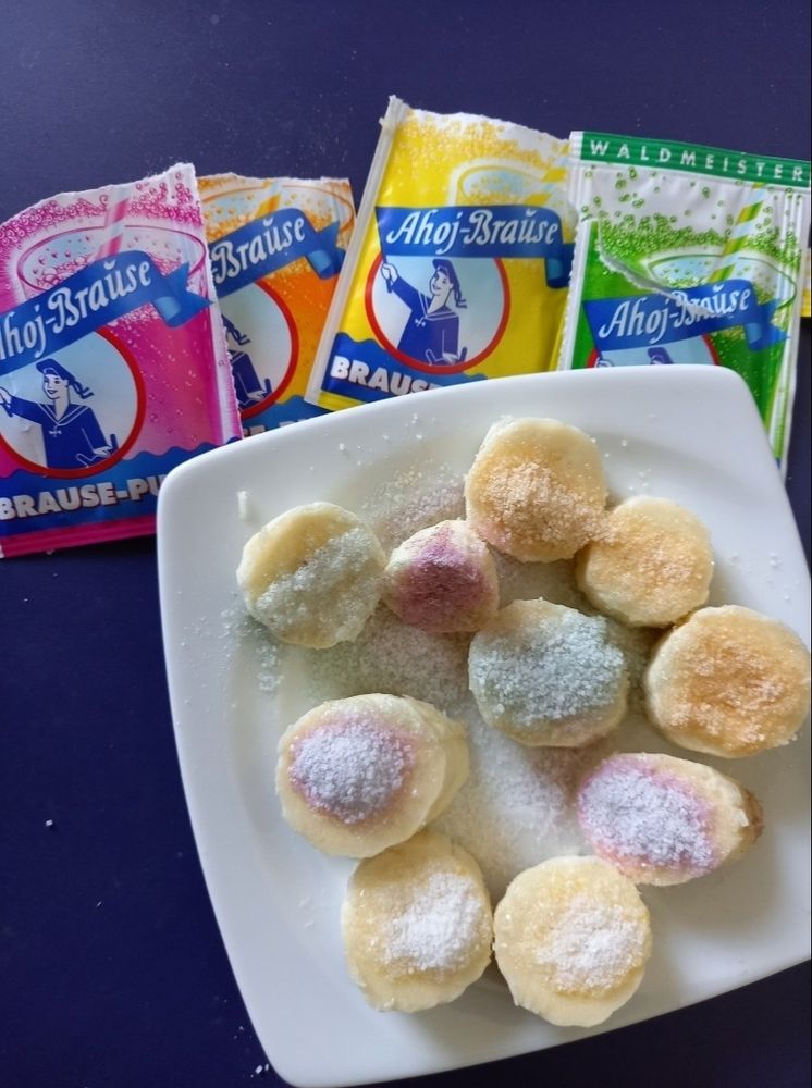 Je 2-3 mit je 3 versch. Ahoj-Brause Pulvern bestreuten Bananenstücke auf weißem quadratischem Teller über 4 aufgerissenen Ahoj-Brause Pulver-Tüten. (Waldmeister-Tüte ungeöffnet) auf Marineblauem Tisch liegend. Von Mobel