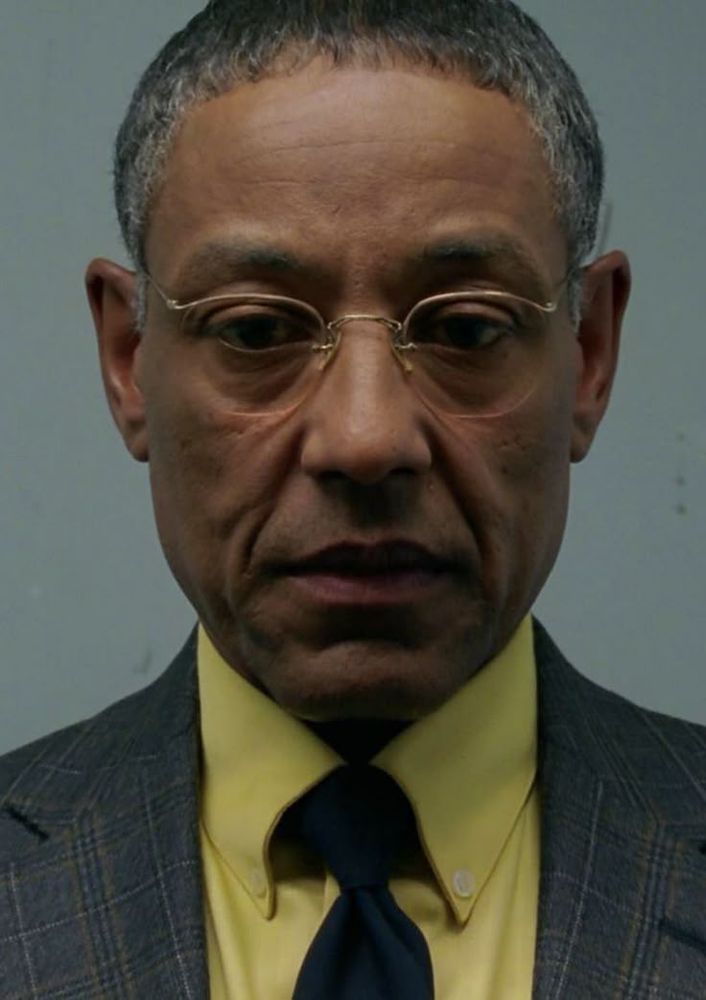 Gus Fring de Breaking Bad, usando óculos e vestindo uma camisa amarela com gravata azul escuro e um paletó, com uma expressão muito séria
