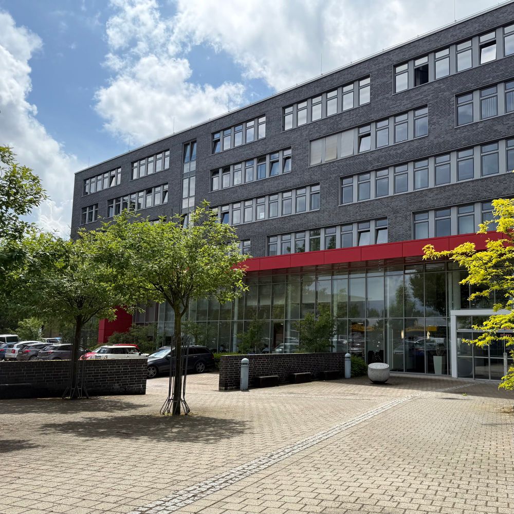 Fassade der Bez.-Reg. Münster mit rotem Eingang und einem Baum auf der linken Seite