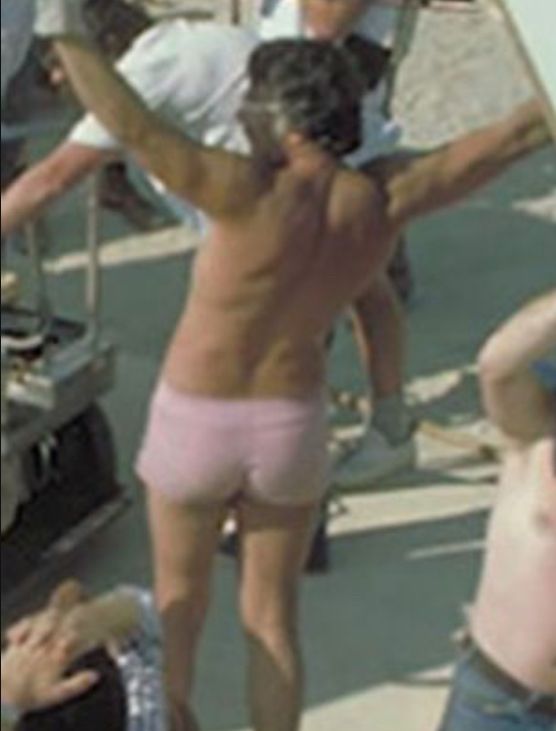 A tan man in Tunisia, holding up a boom (with a mic) that is out of frame.  He is wearing nothing but thick gloves (to hide handling noise) and very short, slutty pink shorts.  His back is to the camera.
