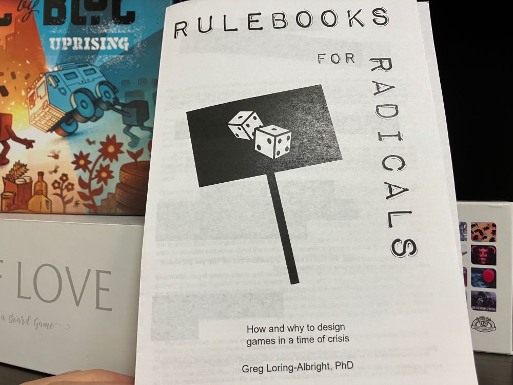 A photo of the cover of the zine, featuring a black protest sign with an icon of rolling dice on it. In the background are some board game boxes, including Bloc by Bloc: Uprising and Fog of Love