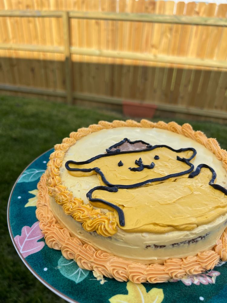A side view of a cake featuring pompompurin holding his arms up. The cake is in a backyard with a fence.