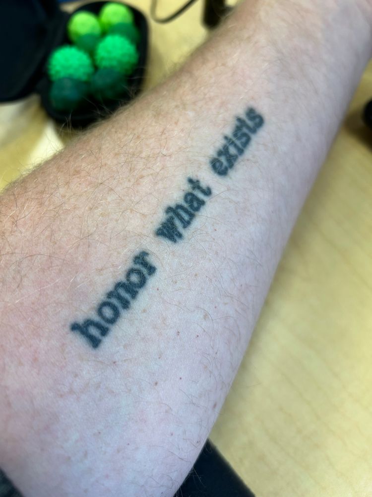 A tattoo on an arm that says in plain text: honor what exists. 