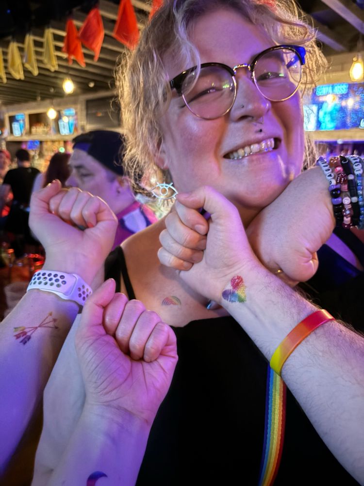 Jamie smiling while people off camera hold up their temporary pride tattoos on their wrists 