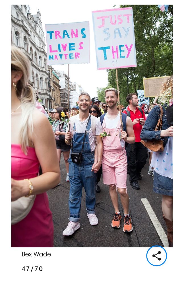 James and Oli holding hands and signs at Trans Pride March. James’ sign says ‘just say they’ and Oli says ‘trans lives matter’ 