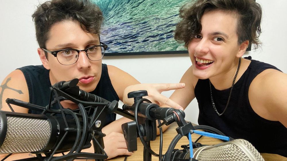 Luc e Fai atrás de microfones em uma mesa, Luc sério, Fai sorrindo e mostrando a língua. Ao fundo, um quadro de onda.