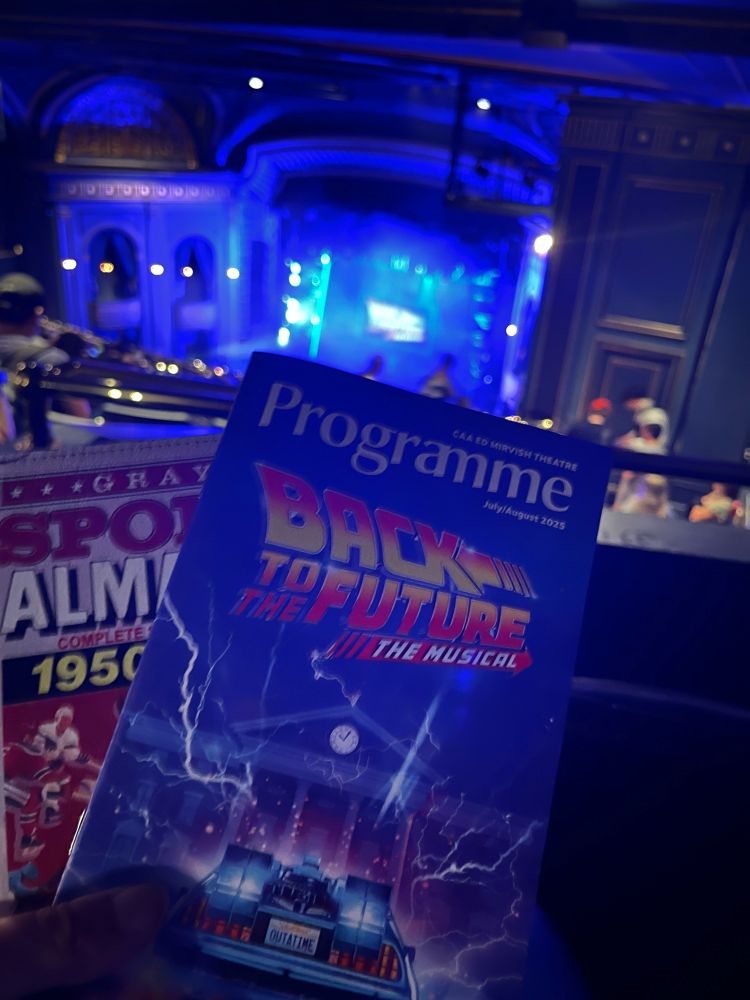 An image of a theatre programme for back to the future musical being held in front of a stage. Also behind the programme is a somewhat obscured copy of Grey’s Sports Almanac (ifykyk)