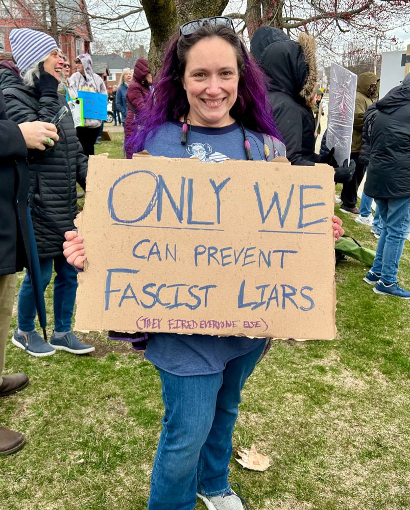 Smiling woman with purple hair standing in a crowd and holding a cardboard sign reading only we can prevent fascist liars (they fired everyone else)