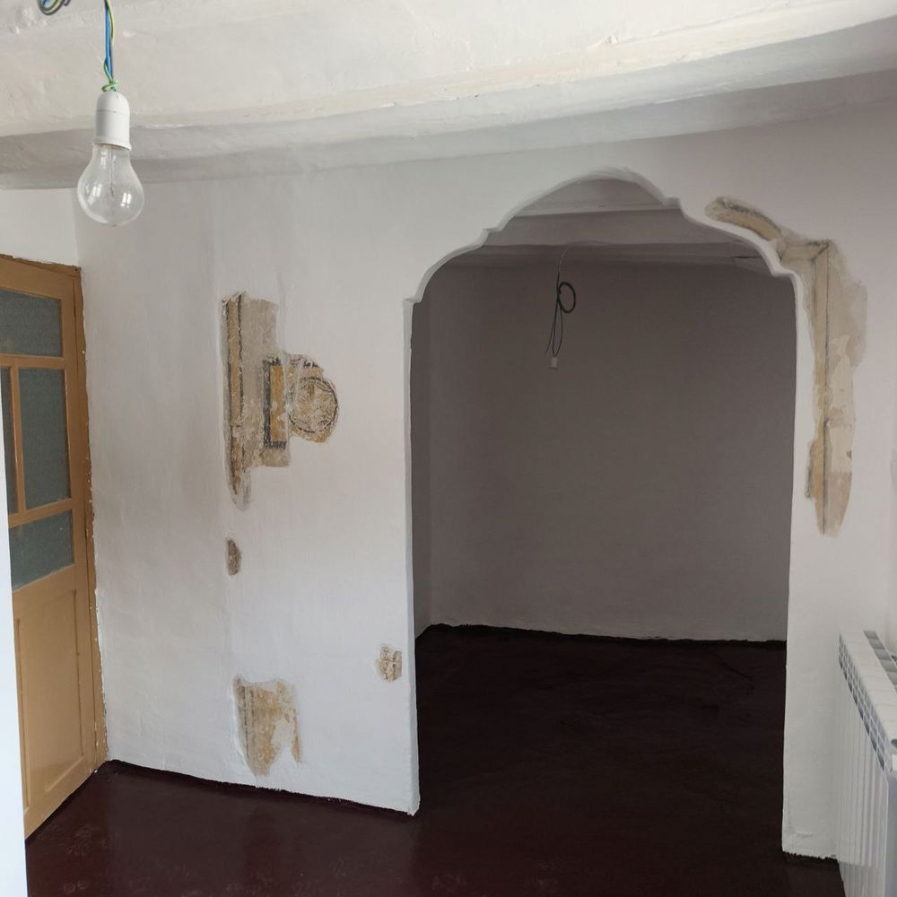 Una habitación antigua, de pueblo, con alcoba. En la pared que da paso a la alcoba se ve un trocito de pintura al fresco. Representa una hexapétala o flor de 6 pétalos. La pared se ha dejado pintada blanca, a la cal. El hueco de entrada a la alcoba tiene unas molduras originales en su parte superior. A la izquierda hay una puerta de madera y cristal, antigua. A la derecha hay un radiador grande, moderno.