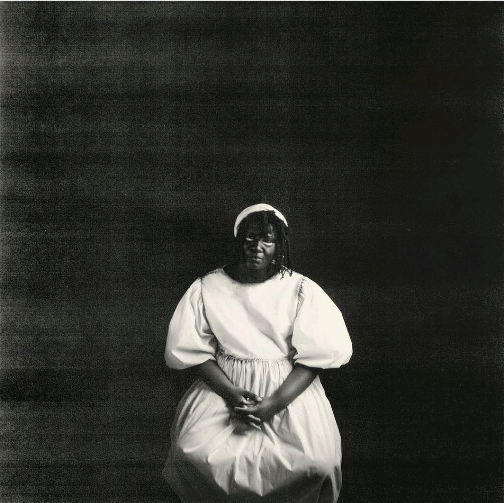 Cover art for Only Dust Remains by Backxwash (pronounced backwash). The artist is a black woman, she wears a puffy white dress and white headband and is sitting down against a grainy black background.