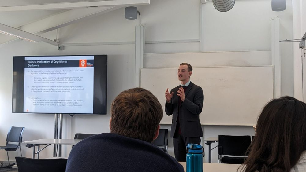 Justin Moll, a white man with dark blonde hair and a moustache. He is wearing a grey suit with a blue shirt. He is stood at the front of a classroom next to a screen showing a slide with the title "Political implications of cognition as disclosure". He is gesturing with his hands as he speaks. The photograph is taken from behind two people's heads.
