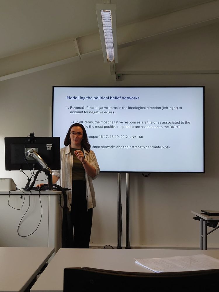 Irene Arahal, a white woman with dark hair and glasses, wearing a light grey shirt over a dark green t shirt and black trousers. She is stood at the front of a classroom in front of a screen showing a slide entitled "Modelling the political belief networks" and is in the middle of speaking, using her hand to highlight a point.