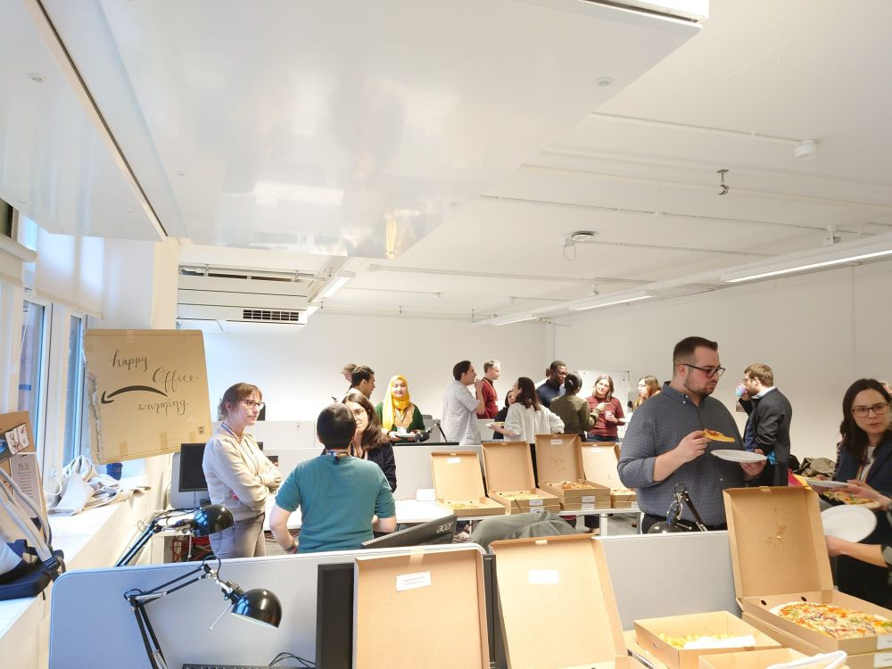 A group of approximately 20 people stood chatting, drinking soft drinks and eating pizza in a brightly lit office space. There are boxes of pizzas open on the tables and a cardboard sign reading "happy office warming"