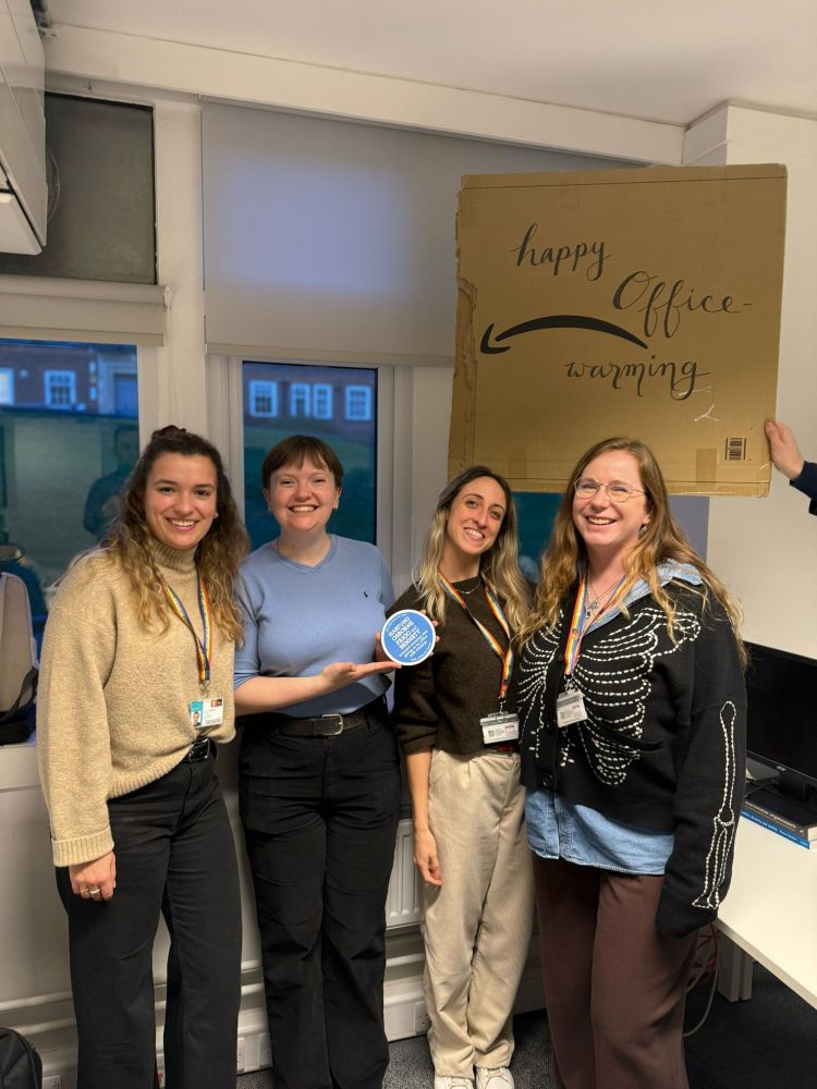 Karia Hartung, Aine Bennett, Lucia Frigo and Kat Osborne, four white women in casual clothes, stand together in an office in front of the windows. They are smiling and have their arms around each other. Aine is holding and gesturing to a small blue plaque. Somebody holds a cardboard sign behind them that says "happy office warming"